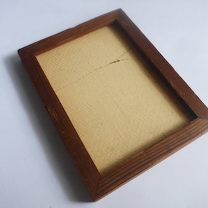 Puede incluir: Un marco de fotos rectangular de madera con acabado marrón oscuro. El marco contiene un respaldo de papel de color beige claro. El marco está vacío y listo para una foto o una obra de arte. El marco está ligeramente inclinado.