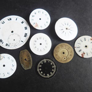 May include: A selection of vintage watch faces in different shapes and sizes. The faces are mostly white with black numerals and hands, some with Roman numerals. One face is gold-coloured, and another is black.
