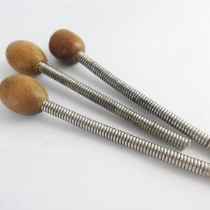 May include: Three wooden-topped, metal-threaded tools are arranged diagonally on a white surface. The wooden tops are oval-shaped and light brown. The metal shafts are silver and have a spiral design.