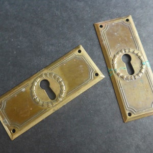 Op de afbeelding: Twee antieke messing sleutelgatbedekkingen met een decoratief kransontwerp. Elke bedekking heeft een sleutelgat in het midden en een rechthoekige vorm.