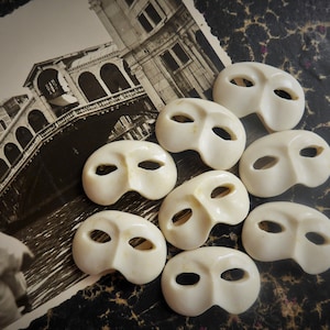 May include: A collection of ten white masquerade masks with eye holes. The masks are arranged in a semi-circle on a textured brown surface.