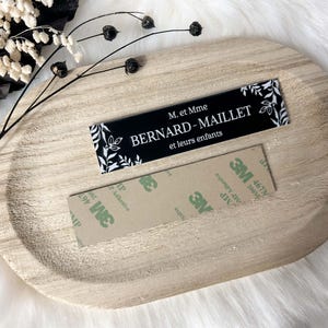 May include: A black and white sign with the text "M. et Mme BERNARD-MAILLET et leurs enfants" and a floral design. The sign is on a wooden surface with adhesive tape.