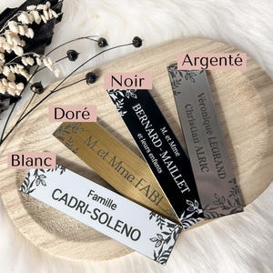 May include: Five different name cards with black lettering on a wooden surface. The cards are in the colors silver, black, gold, white, and a light brown. The text on the cards includes names, titles, and family names.