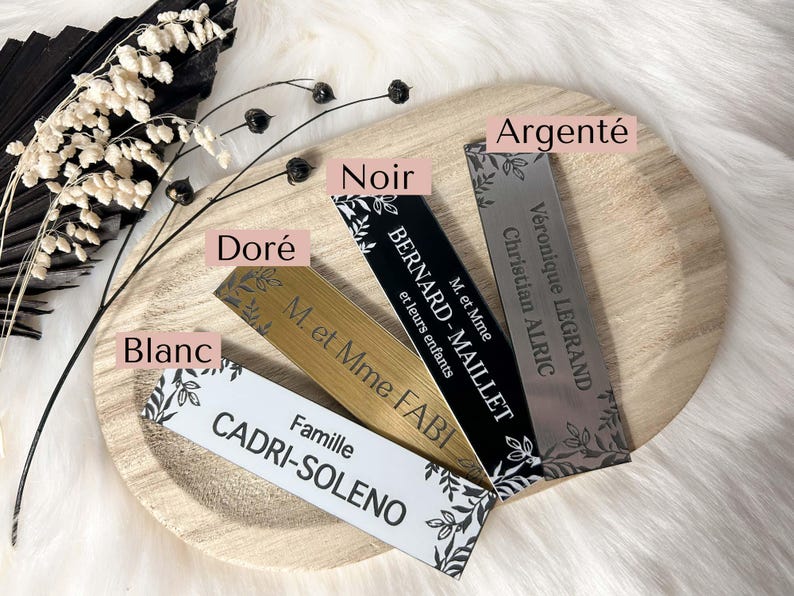 May include: Five different name cards with black lettering on a wooden surface. The cards are in the colors silver, black, gold, white, and a light brown. The text on the cards includes names, titles, and family names.