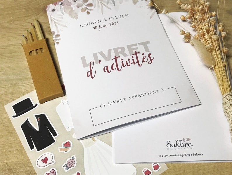 K&ouml;nnte beinhalten: Ein wei&szlig;es Aktivit&auml;tsheft mit dem Titel LIVRET d'activit&eacute;s, den Namen Lauren & Steven und dem Datum 10 juin 2023. Es enth&auml;lt einen Bogen mit Hochzeitsaufklebern, die einen Smoking, ein Kleid und Blumen zeigen, sowie eine kleine Schachtel Buntstifte.