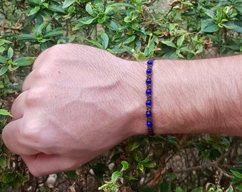 Puede incluir: Una pulsera de macramé con cuentas azul oscuro y un cordón marrón tejido. La pulsera se muestra en una muñeca, con follaje verde de fondo.