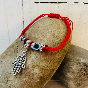 Puede incluir: Pulsera roja tejida con un dije de mano de Hamsa y cuentas decorativas. La pulsera presenta cuentas azules de ojo turco, cuentas facetadas rojas y detalles plateados. El dije de mano de Hamsa es plateado.