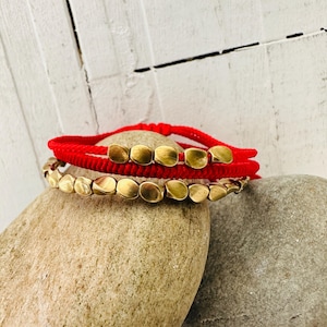 Könnte beinhalten: Zwei Armbänder liegen auf einem Stein. Das obere Armband ist rot mit goldfarbenen quadratischen Perlen. Das untere Armband ist goldfarben mit ovalen Perlen. Der Hintergrund ist eine weiße Holzoberfläche.