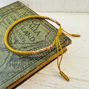 May include: A gold braided cord bracelet with a multi-coloured thread woven into the centre. The bracelet has two long tassels that are tied together with a knot.