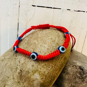 Op de afbeelding: Rode geweven armband met blauwe en witte evil eye kralen. De armband heeft een verstelbare schuifknoopsluiting. De evil eye kralen zijn rond met een blauwe iris en een zwarte pupil.