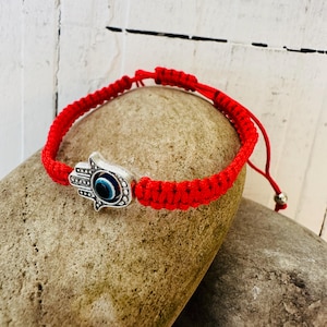 Puede incluir: Pulsera de macramé roja con un dije de mano de Hamsa plateado con un diseño de ojo azul. La pulsera es ajustable y está sobre una piedra. Es un símbolo de protección y buena suerte.