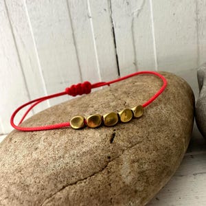 May include: A red cord bracelet featuring five gold-toned, square beads. The bracelet is knotted for size adjustment. The jewellery is presented on a light brown stone, set against a white wooden backdrop.