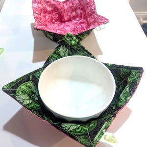 May include: Two fabric bowl cozies. One is pink with a steampunk design, and the other is green with a similar pattern, holding a white bowl. The cozies are square with a star-like shape when open, designed to hold bowls.