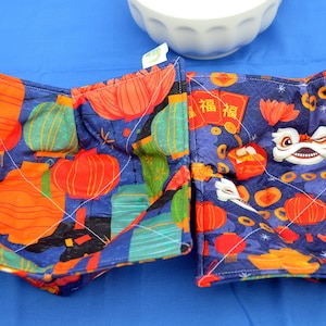 May include: Two blue quilted pot holders with a Chinese New Year theme. One features red lanterns and the other features a lion dance with the Chinese character for "fortune" written on a red envelope.