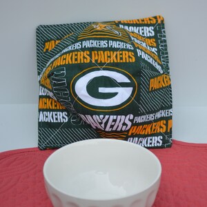 May include: Green and yellow fabric pot holder with the words "Packers" repeated and a green "G" in the center. The pot holder is sitting on a red surface next to a white bowl.