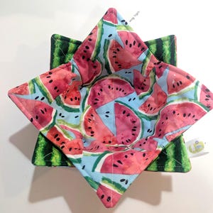 May include: A set of two fabric bowl cozies. The top cozy features a watermelon slice pattern on a light blue background. The bottom cozy has a green watermelon rind pattern. The cozies are square-shaped and designed to hold bowls.