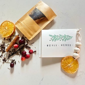 May include: A white greeting card with green pine branch illustrations and the text MERRY MERRY, accompanied by a brown pouch of dried ingredients, a cinnamon stick, dried orange slices, red berries, cloves, and three white wooden beads.