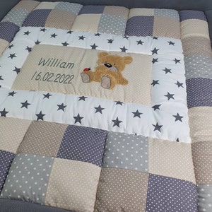 Könnte beinhalten: Eine weiße und beige Patchwork-Babydecke mit grauem Rand. Die Decke hat ein zentrales Feld mit einem braunen Teddybär und dem Text "William 16.02.2022".