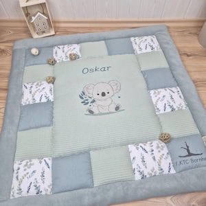 Può includere: Tappeto gioco per bambini con design patchwork nei toni del blu e del verde. Il centro presenta un'illustrazione di un koala e il nome "Oskar". Elementi decorativi includono motivi floreali e piccole palline di legno.