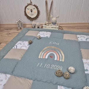Może przedstawiać: Niebieska i beżowa mata do zabawy w patchworku z wyszytym tęczą i imieniem "Ella". Data "11.10.2024" również jest wyszyta na macie.