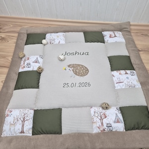 May include: A personalized baby blanket with a patchwork design. The center features the name "Joshua", a hedgehog illustration, and the date "25.01.2026". The blanket combines olive green, beige, and patterned fabric squares, framed by a brown border.