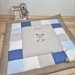 Può includere: Un tappetino da gioco patchwork quadrato con un pannello centrale grigio chiaro a trama waffle, ricamato con il nome Theo e un piccolo disegno di un orsetto. Il bordo è composto da quadrati di tessuto alternati blu, azzurri, bianchi e grigi.