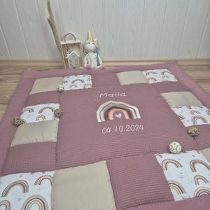 May include: A mauve baby play mat with a patchwork design. The center features a stitched rainbow and the name "Maila" with the date "04.10.2024". The mat is surrounded by beige and patterned squares with rainbow motifs. A unicorn plush toy and decorative items are in the background.