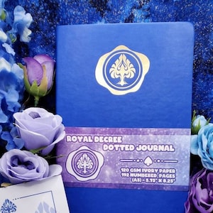 Puede incluir: Un diario azul con un diseño de pica dorado y el texto "Royal Decree Dotted Journal" en la portada. El diario es de papel marfil de 120 GSM con 192 páginas numeradas y mide 14,6 cm x 21 cm.