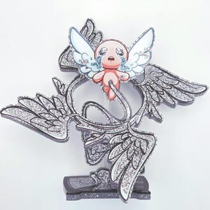 May include: A glittery, stylized angel figurine with a crying baby figure at the center. The baby has white wings and is surrounded by a silver, wing-shaped design. The figurine is mounted on a black base.