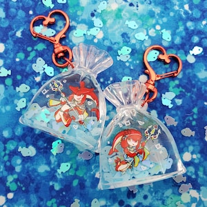 May include: Two clear plastic bag charms with red heart-shaped keychains. Each bag contains a red-haired character with a blue and white outfit holding a trident. The bags are filled with blue and white glitter.