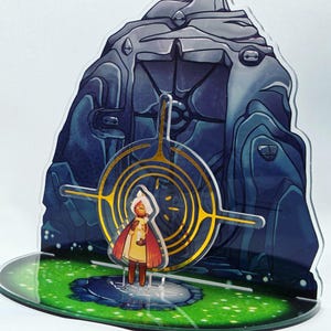 May include: A 3D printed acrylic figurine of a character from the video game "Ori and the Will of the Wisps". The character is standing in front of a large, blue, rocky structure with a golden circle around it. The base of the figurine is green and has a small puddle of water on it.