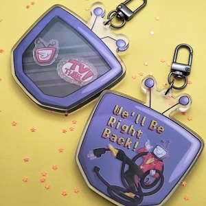 Puede incluir: Dos llaveros acrílicos con forma de TV con un diseño de dibujos animados. Un llavero tiene el texto "TV TIME!" y un personaje de dibujos animados de TV. El otro llavero tiene el texto "We'll Be Right Back!" y un personaje de dibujos animados de TV.