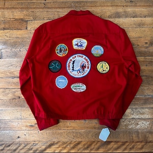 Könnte beinhalten: Eine leuchtend rote Langarmjacke mit einem großen, runden Aufnäher des Order of the Arrow Boy Scouts of America in der Mitte, umgeben von sieben kleineren gestickten Abzeichen, darunter Klondike Derby und District Camporee Motive.