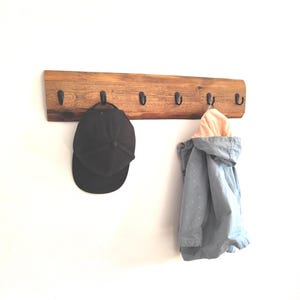 Könnte beinhalten: Ein hölzerner Wand-Kleiderhaken mit sechs schwarzen Metallhaken. Eine schwarze Baseballkappe hängt an einem Haken, und eine hellblaue Jacke mit rosa Futter hängt an einem anderen. Das Holz hat eine natürliche, rustikale Oberfläche.