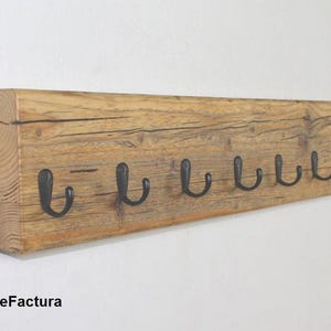 Könnte beinhalten: Ein rustikaler Garderobenständer aus Holz mit sieben schwarzen Metallhaken. Das Holz hat ein natürliches, verwittertes Aussehen mit sichtbarer Maserung und Knoten. Die Haken sind gleichmäßig am unteren Rand des Holzbretts angeordnet.