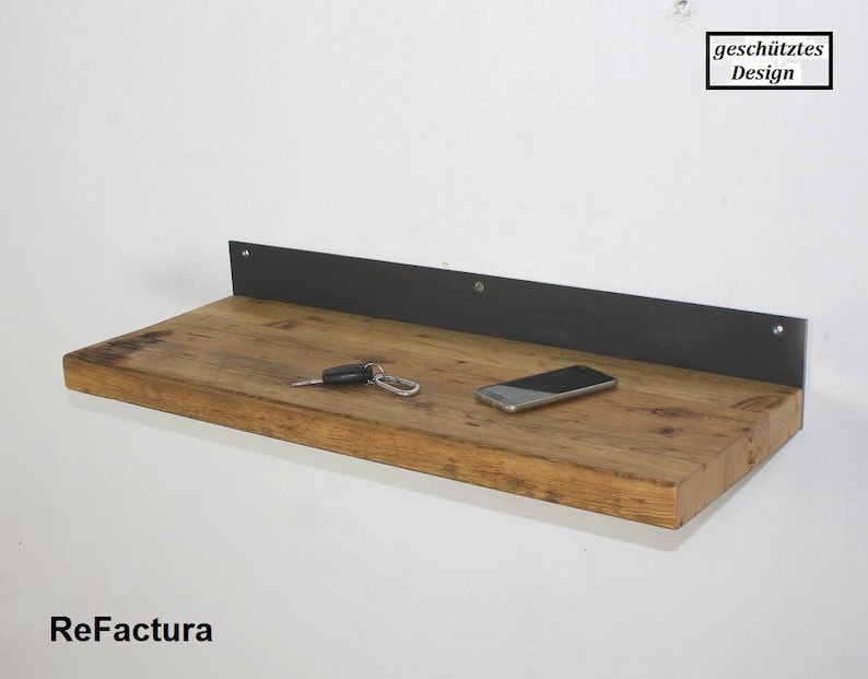 May include: A rustic wooden shelf with a black metal bracket. The shelf is mounted to a white wall and has a cell phone and keys on it. The text "gesch&uuml;tztes Design" is visible in the upper right corner of the image.