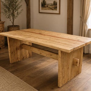 Könnte beinhalten: Ein großer, rechteckiger Esstisch aus Holz mit hellbraunem Finish. Der Tisch hat dicke Beine und einen Stützbalken. Die Tischplatte hat einen rotbraunen Akzentstreifen. Der Tisch steht in einem hellen Raum.