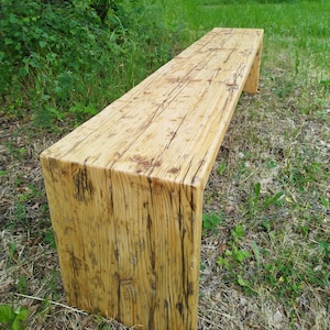 Pu&ograve; includere: Panca in legno rustico con finitura naturale. La panca rettangolare ha una seduta spessa e gambe a blocco. Le venature del legno e i nodi sono visibili, conferendole un aspetto invecchiato. La panca &egrave; posizionata sull'erba.