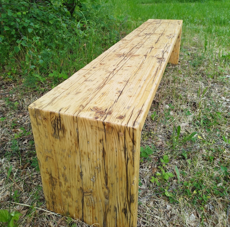Pu&ograve; includere: Panca in legno rustico con finitura naturale. La panca rettangolare ha una seduta spessa e gambe a blocco. Le venature del legno e i nodi sono visibili, conferendole un aspetto invecchiato. La panca &egrave; posizionata sull'erba.