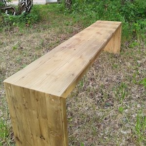 Pu&ograve; includere: Panca in legno chiaro con seduta rettangolare e gambe spesse a forma di blocco. La panca &egrave; realizzata in legno naturale con venature e nodi visibili. &Egrave; all'aperto, su erba e circondata da alberi.