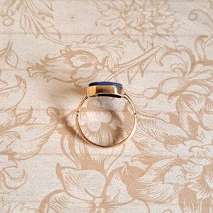 K&ouml;nnte beinhalten: Ein goldener Ring mit einem dunkelblauen Edelstein. Der Ring liegt auf einem floralen Hintergrund.