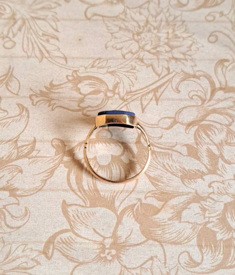 K&ouml;nnte beinhalten: Ein goldener Ring mit einem dunkelblauen Edelstein. Der Ring liegt auf einem floralen Hintergrund.