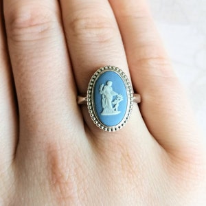 May include: A silver ring with a blue oval cameo featuring a white ceramic figure of a woman holding a flower.