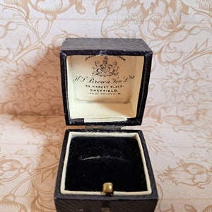 K&ouml;nnte beinhalten: Eine ge&ouml;ffnete schwarze Ringbox mit wei&szlig;em Innenraum. Die Box ist mit schwarzem Samt ausgekleidet und hat ein goldenes Scharnier. Die Box ist mit "H.P. Brown Jon. Ltd. 65 Market Place, Sheffield. And at London, W." beschriftet.