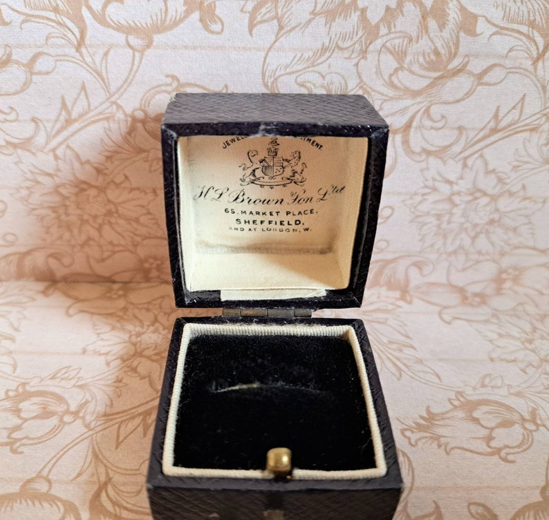 K&ouml;nnte beinhalten: Eine ge&ouml;ffnete schwarze Ringbox mit wei&szlig;em Innenraum. Die Box ist mit schwarzem Samt ausgekleidet und hat ein goldenes Scharnier. Die Box ist mit "H.P. Brown Jon. Ltd. 65 Market Place, Sheffield. And at London, W." beschriftet.
