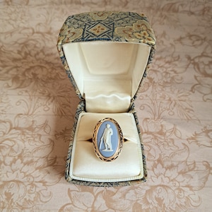 Pu&ograve; includere: Un anello cammeo blu con una montatura in oro, che presenta un cammeo bianco di una donna con un vestito lungo, in una piccola scatola di velluto.