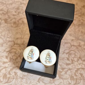 May include: A pair of round cufflinks featuring a Peter Rabbit illustration on a white background with gold-tone rims, presented inside an open black rectangular box with a black velvet interior lining.