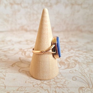 K&ouml;nnte beinhalten: Ein goldener Ring mit einem blauen Edelstein, der in einer Zargenfassung gefasst ist. Der Ring befindet sich auf einem h&ouml;lzernen Ringst&auml;nder.