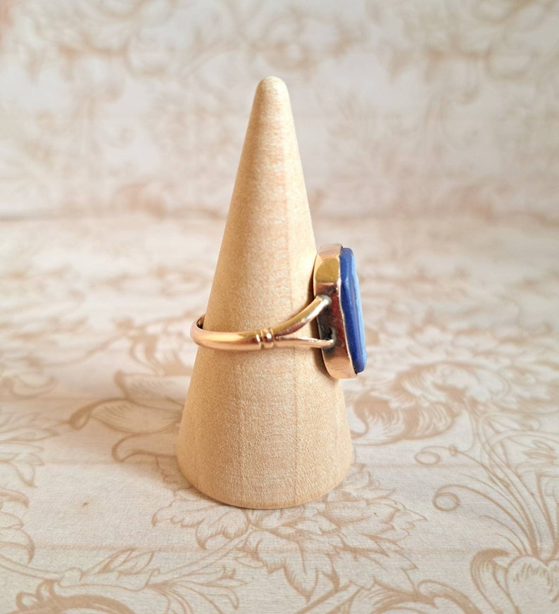 K&ouml;nnte beinhalten: Ein goldener Ring mit einem blauen Edelstein, der in einer Zargenfassung gefasst ist. Der Ring befindet sich auf einem h&ouml;lzernen Ringst&auml;nder.