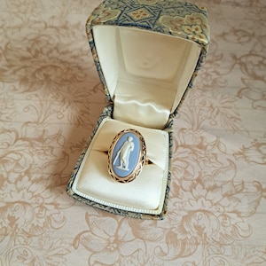 Pu&ograve; includere: Un anello con cammeo blu e bianco in una montatura dorata, esposto in una scatola con motivo.